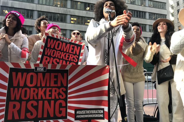 A group of women sing at a rally pushing to increase the minimum wage for restaurant workers carrying signs that say Women Workers Rising.